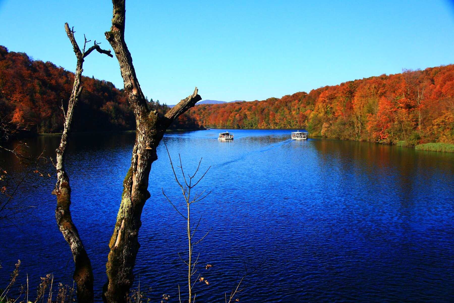 Lac de Plitvice d’un bleu profond bordé de forêts rousses et dorées en octobre, deux bateaux électriques au loin